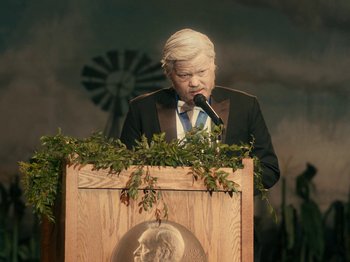 Movie still from “I'm Thinking of Ending Things” (2020), directed by Charlie Kaufman – An older man in a suit is speaking into a microphone; Medium shot, Over the shoulder angle