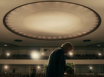 Movie still from “I'm Thinking of Ending Things” (2020), directed by Charlie Kaufman – A man standing in front of an audience in front of a stage; Wide shot, Low angle