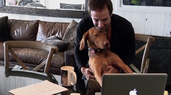 Movie still from “Icarus” (2017), directed by Bryan Fogel – A man sitting at a table with a dog and a laptop; Medium shot, Low angle