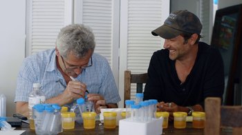 Movie still from “Icarus” (2017), directed by Bryan Fogel – Two men sitting at a wooden table with cups of juice; Medium shot, Over the shoulder angle