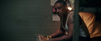 Movie still from “Kaleidoscope” (2023), directed by Mairzee Almas – A man sitting in front of a brick wall holding a magazine; Medium shot, Over the shoulder angle