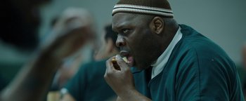 Movie still from “Kaleidoscope” (2023), directed by Mairzee Almas – A man eating a donut in a room with others; Close Up shot, Over the shoulder angle
