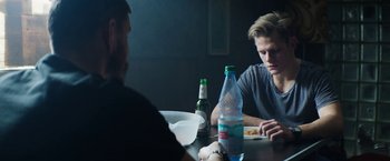 Movie still from “Kidnapping Stella” (2019), directed by Thomas Sieben – Two men sitting at a table with a bottle of beer and a plate; Medium shot, Over the shoulder angle
