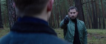Movie still from “Kidnapping Stella” (2019), directed by Thomas Sieben – A man holding a gun pointed at another man in the woods; Medium shot, Over the shoulder angle
