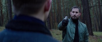 Movie still from “Kidnapping Stella” (2019), directed by Thomas Sieben – A man holding a gun in his hand while standing next to another man in the woods; Medium shot, Over the shoulder angle
