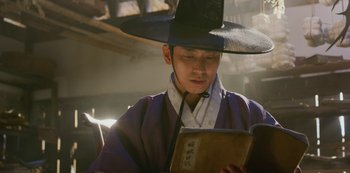 Movie still from “Kingdom” (2019), created by Eun-hee Kim – A man wearing a hat and holding an open book; Close Up shot, Low angle