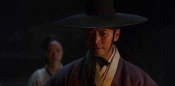 Movie still from “Kingdom” (2019), created by Eun-hee Kim – A man wearing a black hat standing next to a woman; Close Up shot, Over the shoulder angle