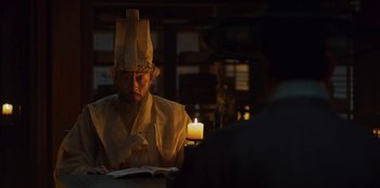 Movie still from “Kingdom” (2019), created by Eun-hee Kim – A man wearing a paper crown sitting at a table; Medium shot, Over the shoulder angle