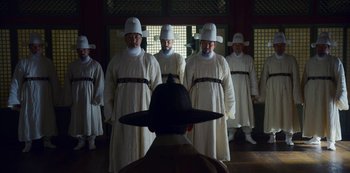 Movie still from “Kingdom” (2019), created by Eun-hee Kim – A group of men in white robes and white hats standing next to each other; Wide shot, Over the shoulder angle
