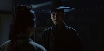 Movie still from “Kingdom” (2019), created by Eun-hee Kim – A man wearing a black hat standing next to a woman; Close Up shot, Over the shoulder angle