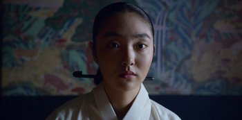 Movie still from “Kingdom” (2019), created by Eun-hee Kim – A young woman wearing a white shirt and a white shirt with a wooden handle on her head; Close Up shot, Low angle