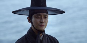 Movie still from “Kingdom” (2019), created by Eun-hee Kim – A man wearing a black hat and a neck tie; Close Up shot, Low angle