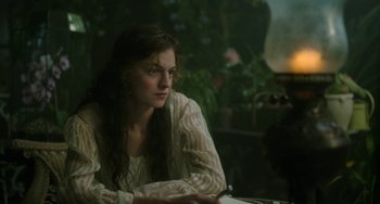 Movie still from “Lady Chatterley's Lover” (2022), directed by Laure de Clermont-Tonnerre – A woman sitting at a table in front of a plant; Medium shot, Over the shoulder angle