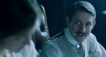 Movie still from “Lady Chatterley's Lover” (2022), directed by Laure de Clermont-Tonnerre – A man sitting in a chair wearing a suit and tie; Close Up shot, Over the shoulder angle