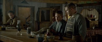 Movie still from “Lawless” (2012), directed by John Hillcoat – Two men sitting at a wooden table in front of a bottle; Medium shot, Over the shoulder angle