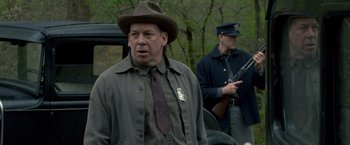 Movie still from “Lawless” (2012), directed by John Hillcoat – A man wearing a hat and a tie standing next to another man holding a gun; Medium shot, Over the shoulder angle