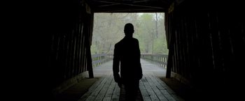 Movie still from “Lawless” (2012), directed by John Hillcoat – A man standing on a wooden bridge with trees in the background; Wide shot, Low angle