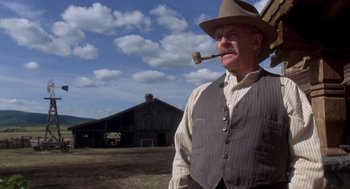 Movie still from “Legends of the Fall” (1994), directed by Edward Zwick – An older man wearing a hat and holding a pipe in his mouth while standing in front of an old barn; Medium shot, Low angle