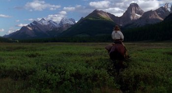 Movie still from “Legends of the Fall” (1994), directed by Edward Zwick – A man riding a horse through a field with mountains in the background; Wide shot, Low angle