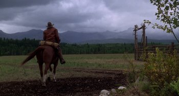 Movie still from “Legends of the Fall” (1994), directed by Edward Zwick – A man riding a horse through a field with mountains in the background; Wide shot, Low angle