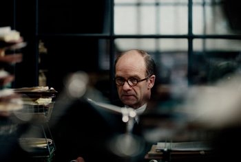 Movie still from “Living” (2022), directed by Oliver Hermanus – A man sitting in front of a laptop computer; Medium shot, Over the shoulder angle