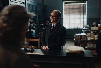 Movie still from “Living” (2022), directed by Oliver Hermanus – A man sitting at a desk in a suit and tie; Medium shot, Over the shoulder angle