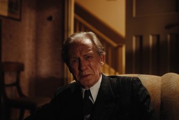 Movie still from “Living” (2022), directed by Oliver Hermanus – An older man wearing a suit and tie sitting in a chair; Medium shot, High angle
