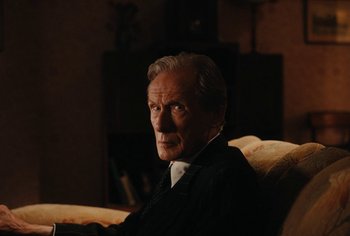 Movie still from “Living” (2022), directed by Oliver Hermanus – An older man sitting on a couch in a room; Close Up shot, Over the shoulder angle