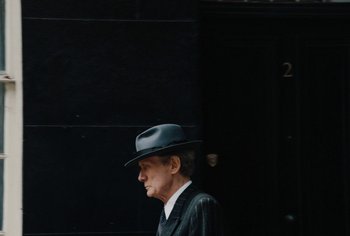 Movie still from “Living” (2022), directed by Oliver Hermanus – A man wearing a hat walking down the street; Medium shot, Over the shoulder angle