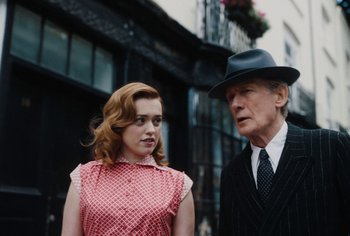 Movie still from “Living” (2022), directed by Oliver Hermanus – An older man and a young woman standing next to each other on the street; Medium shot, Over the shoulder angle