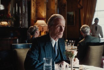 Movie still from “Living” (2022), directed by Oliver Hermanus – An older man sitting at a table in front of a group of people; Medium shot, Over the shoulder angle