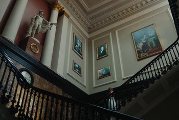 Movie still from “Living” (2022), directed by Oliver Hermanus – A staircase in a large room with paintings on the walls; Extreme Wide shot, Low angle
