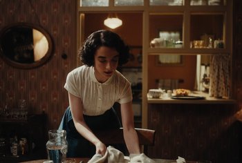 Movie still from “Living” (2022), directed by Oliver Hermanus – A woman in white shirt and black skirt cleaning a table; Medium shot, Over the shoulder angle
