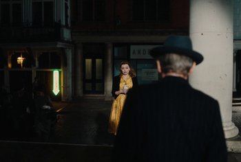 Movie still from “Living” (2022), directed by Oliver Hermanus – A woman in a yellow dress standing in front of a man in a black suit; Wide shot, Over the shoulder angle