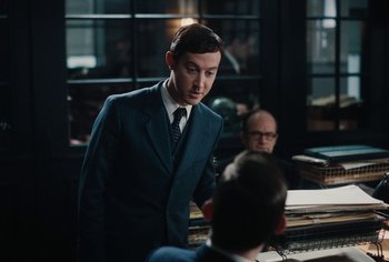 Movie still from “Living” (2022), directed by Oliver Hermanus – A man wearing a suit and tie standing in front of a piano; Medium shot, Over the shoulder angle