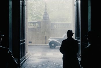 Movie still from “Living” (2022), directed by Oliver Hermanus – A man in a hat standing in front of an open gate; Medium shot, Over the shoulder angle