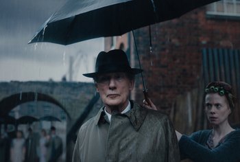 Movie still from “Living” (2022), directed by Oliver Hermanus – An older man in a hat and coat holding an umbrella; Medium shot, Over the shoulder angle