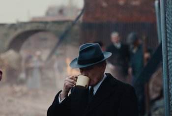 Movie still from “Living” (2022), directed by Oliver Hermanus – A man in a suit and hat drinking from a cup; Medium shot, Over the shoulder angle
