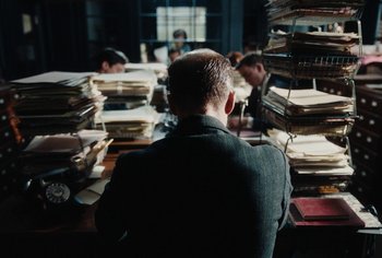 Movie still from “Living” (2022), directed by Oliver Hermanus – A man sitting in front of a stack of papers; Medium shot, Over the shoulder angle