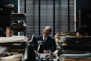 Movie still from “Living” (2022), directed by Oliver Hermanus – An older man sitting at a desk in front of stacks of papers; Medium shot, High angle