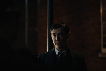 Movie still from “Living” (2022), directed by Oliver Hermanus – A man in a suit standing in a dark room; Close Up shot, Over the shoulder angle