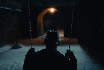 Movie still from “Living” (2022), directed by Oliver Hermanus – A man wearing a hat is sitting on a swing at night; Wide shot, Over the shoulder angle