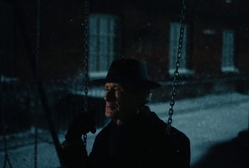 Movie still from “Living” (2022), directed by Oliver Hermanus – An old man wearing a hat is on a swing in the dark; Close Up shot, Low angle
