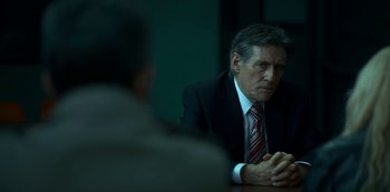 Movie still from “Lost Girls” (2020), directed by Liz Garbus – A man in a suit and tie sitting at a table; Medium shot, Over the shoulder angle