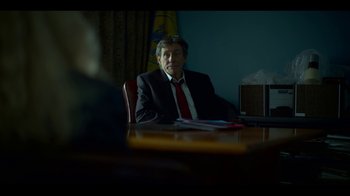 Movie still from “Lost Girls” (2020), directed by Liz Garbus – A man sitting at a table in a suit and tie; Medium shot, Over the shoulder angle