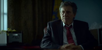 Movie still from “Lost Girls” (2020), directed by Liz Garbus – A man in a suit and tie sitting in front of a curtain; Close Up shot, Low angle