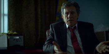 Movie still from “Lost Girls” (2020), directed by Liz Garbus – An older man wearing a suit and red tie sitting at a table; Close Up shot, Over the shoulder angle