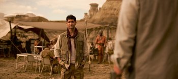 Movie still from “Lost in Space” (2018), created by Irwin Allen – A man standing in the dirt with other people in the background; Medium shot, Over the shoulder angle