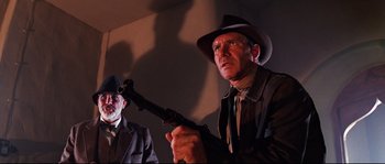 Movie still from “Indiana Jones and the Last Crusade” (1989), directed by Steven Spielberg – A man holding a gun while standing next to another man; Medium shot, Low angle