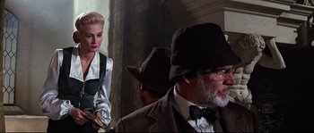 Movie still from “Indiana Jones and the Last Crusade” (1989), directed by Steven Spielberg – A man and a woman in a room with a man wearing a suit and a hat; Medium shot, Over the shoulder angle